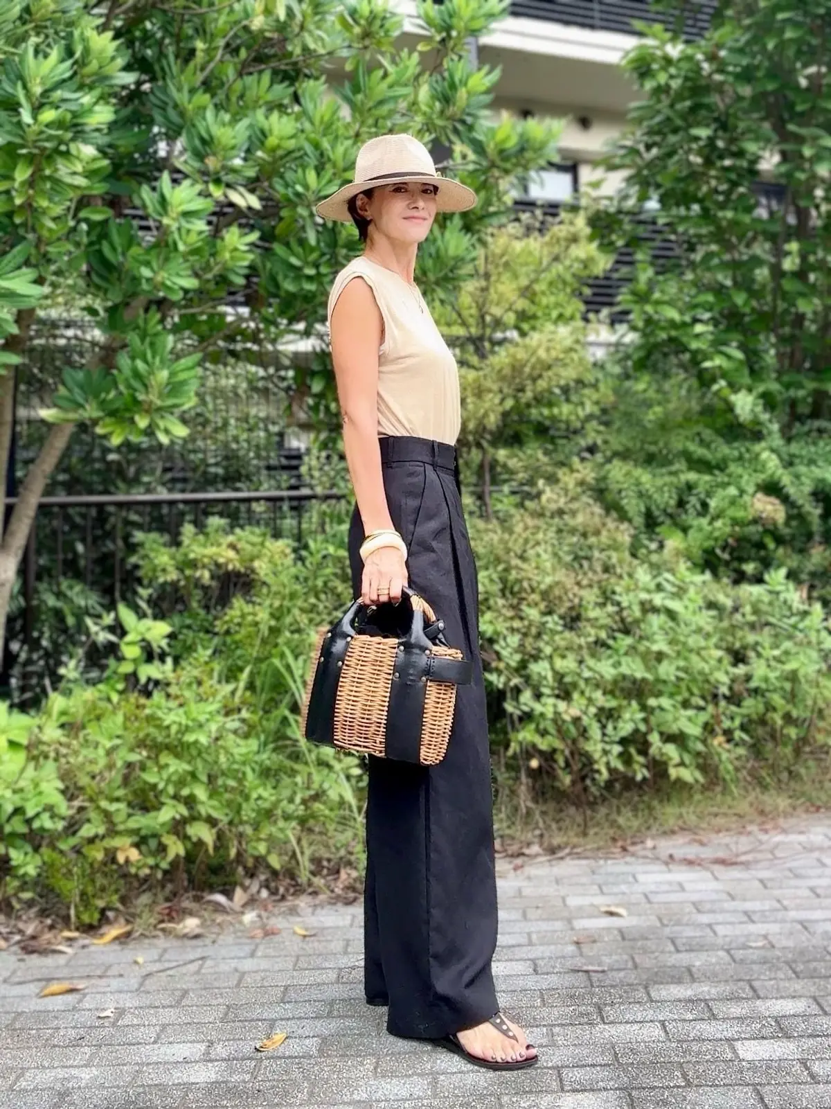 ベージュの帽子を被った女性が、黒いワイドパンツとタンクトップ姿で立っている全身写真。右手に編み込みのバッグを持ち、背景には緑の植物が生い茂っている。