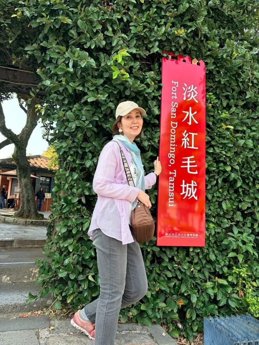 濃い緑の低木に囲まれ、赤い看板を持つ女性が帽子とスカーフを着用し、笑顔で立っている
