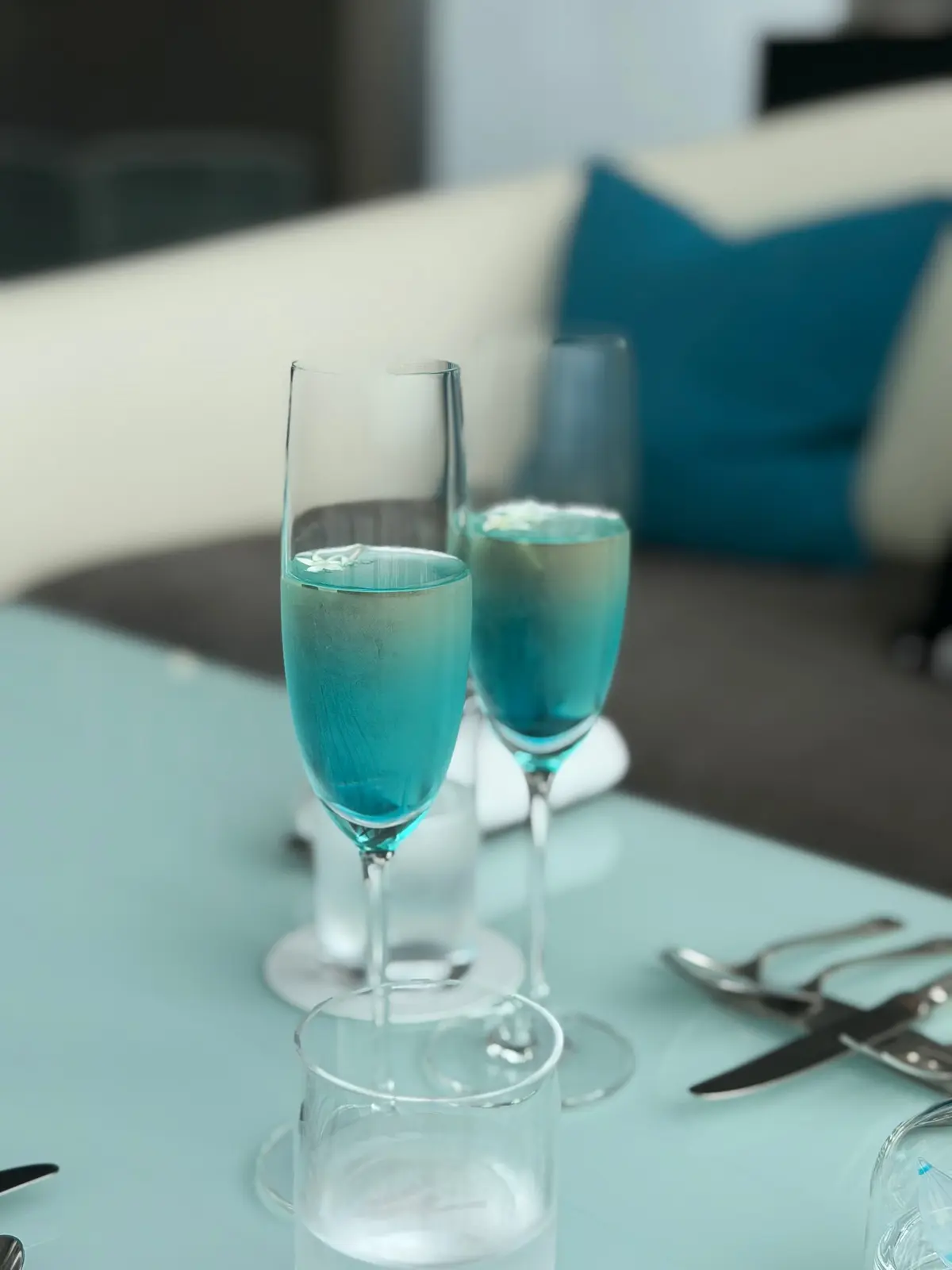 青いテーブルに青いカクテルが２杯と水のグラスが１杯、ナイフとスプーンが置かれている