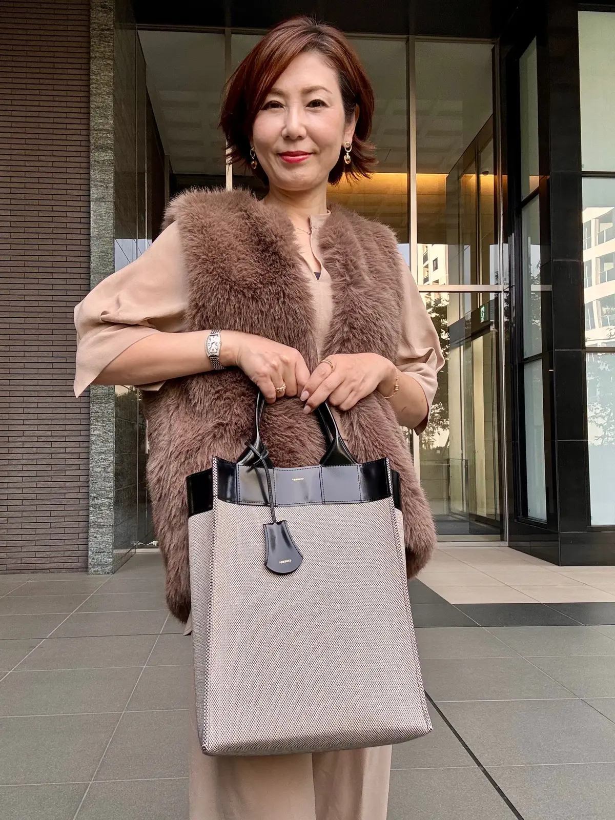 ベージュのパンツとシャツに、茶色の毛皮ベストを着た女性。右手にデルヴォーのトートバッグを持ち、背景に近代的な建物の入り口がある。金色のイヤリングと腕時計を身に着けている。