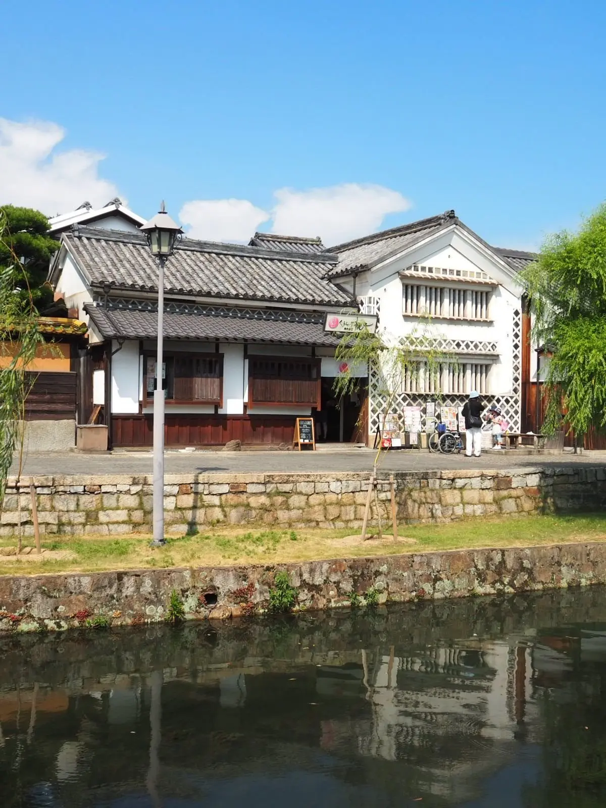 水辺に面した、白い壁と黒い屋根の伝統的な日本の建物。石垣と草地の縁取りがあり、建物は水面に反射している。青空に白い雲が浮かんでいる。