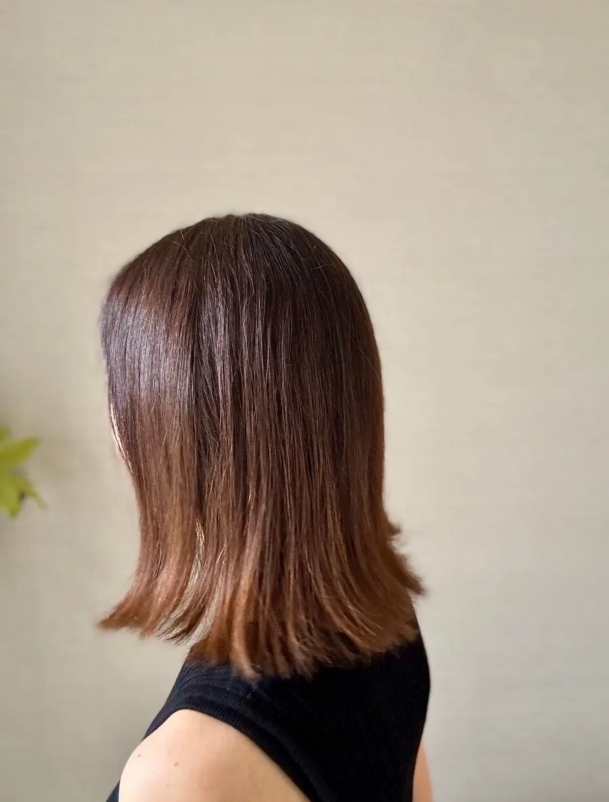 茶色のボブヘアを肩まで伸ばした女性の後ろ姿。シンプルな黒いタンクトップを着用。背景はベージュ色の壁。