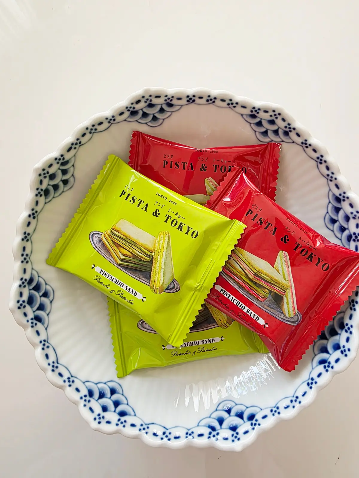 ピスタチオサンドの個包装が4つ、青と白の模様の皿に置かれている。緑と赤のパッケージで、それぞれ「PISTA & TOKYO」のロゴが印刷されている。