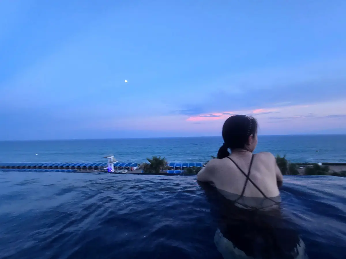 プールサイドで海を眺める女性。水面は青く、遠くには夕焼け空と月が見える。背景には海岸線と緑の植栽が広がる。