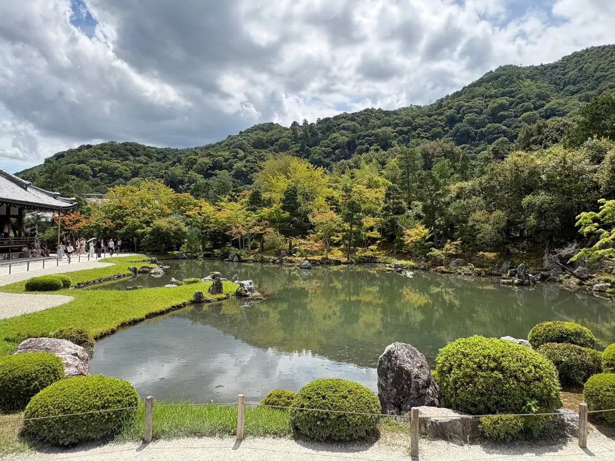 池と緑豊かな山を背景にした庭園。手前に丸い植栽が並び、女性がスマートフォンで撮影している。水面には空の雲の反射が見える。