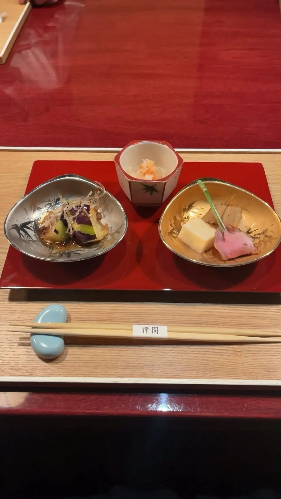 赤い台に3つの料理が並ぶ。左は銀の椀に野菜と海藻、中央は白い小鉢にオレンジ色の具、右は金色の椀に豆腐と花のついた魚の切り身。木製のテーブルに青い石の置物と「洋風」の文字が入った箸が置かれている。