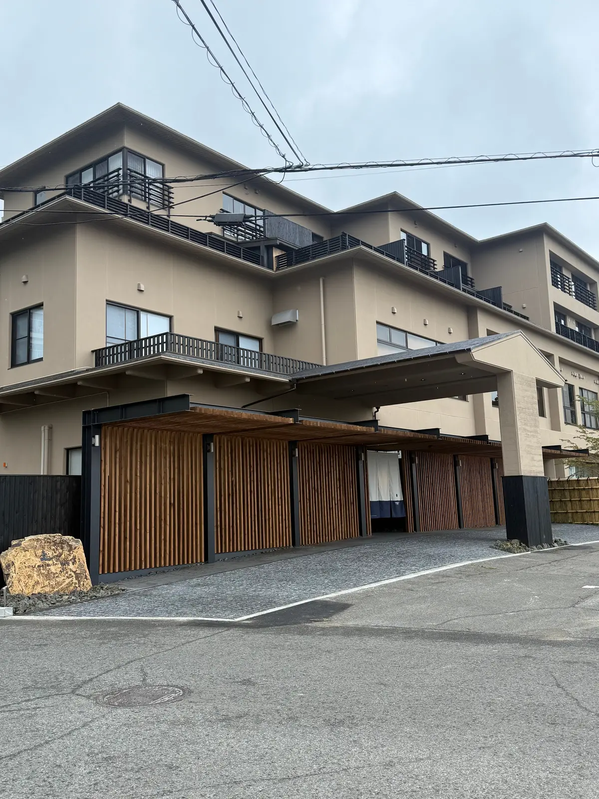ベージュ色の多層住宅と、木製のルーバー塀で囲まれた駐車場。屋根付きの駐車場は、住宅の前に位置し、道路に面している。空は曇り。