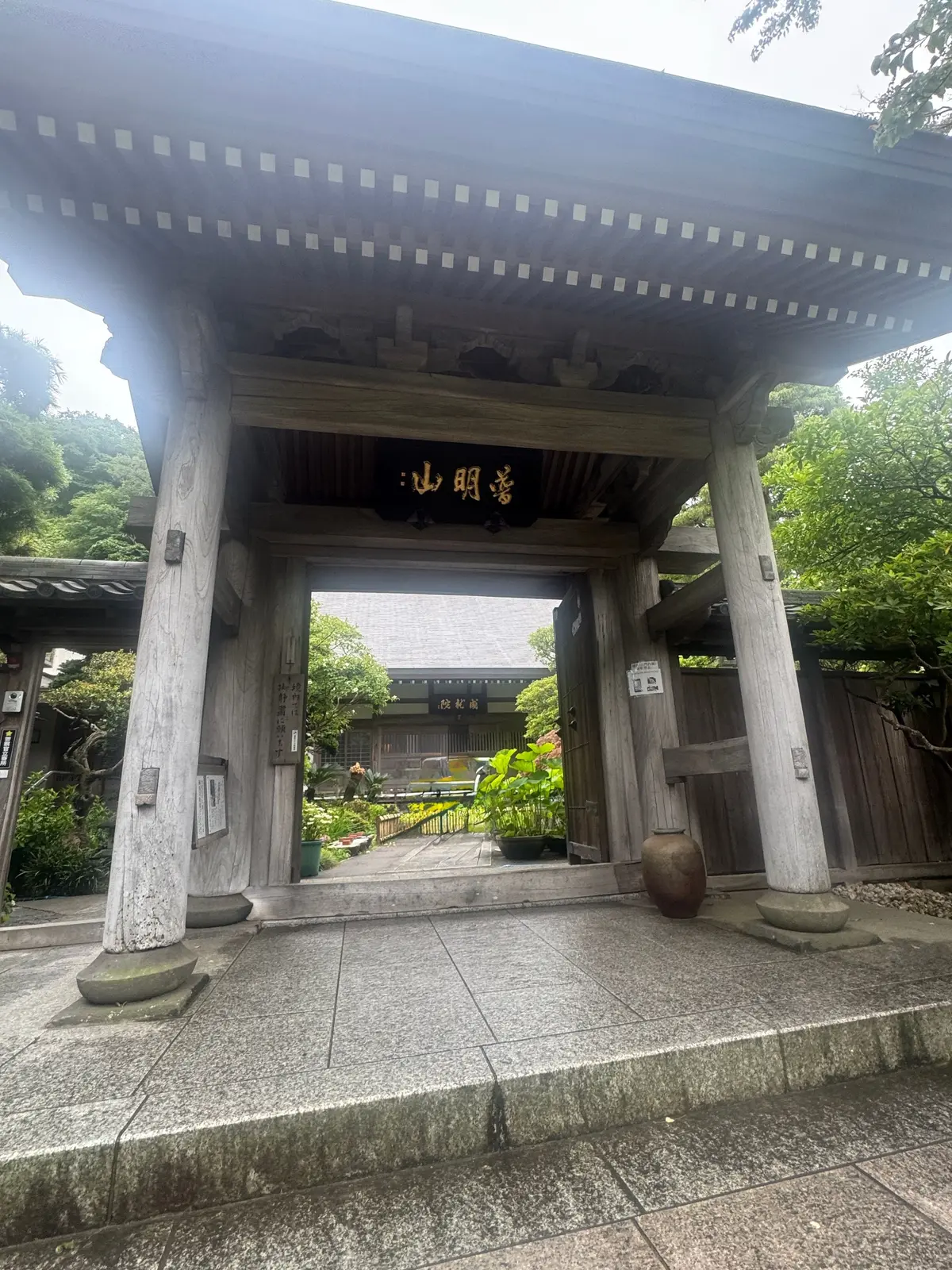 木造の門が石の床に立ち、中央に「心明寺」の文字が掲げられている。門柱に植えられた緑の植物と、右側に置かれた陶器の壺が特徴的。背景には木造の建物と緑豊かな木々が広がる。