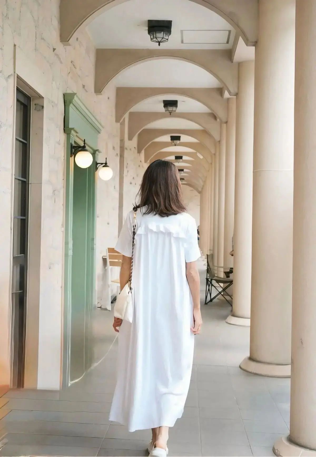 白いワンピースを着た女性がアーチ型の柱が並ぶ通路を歩く。壁には丸い照明が取り付けられ、左側に緑のドアがある。