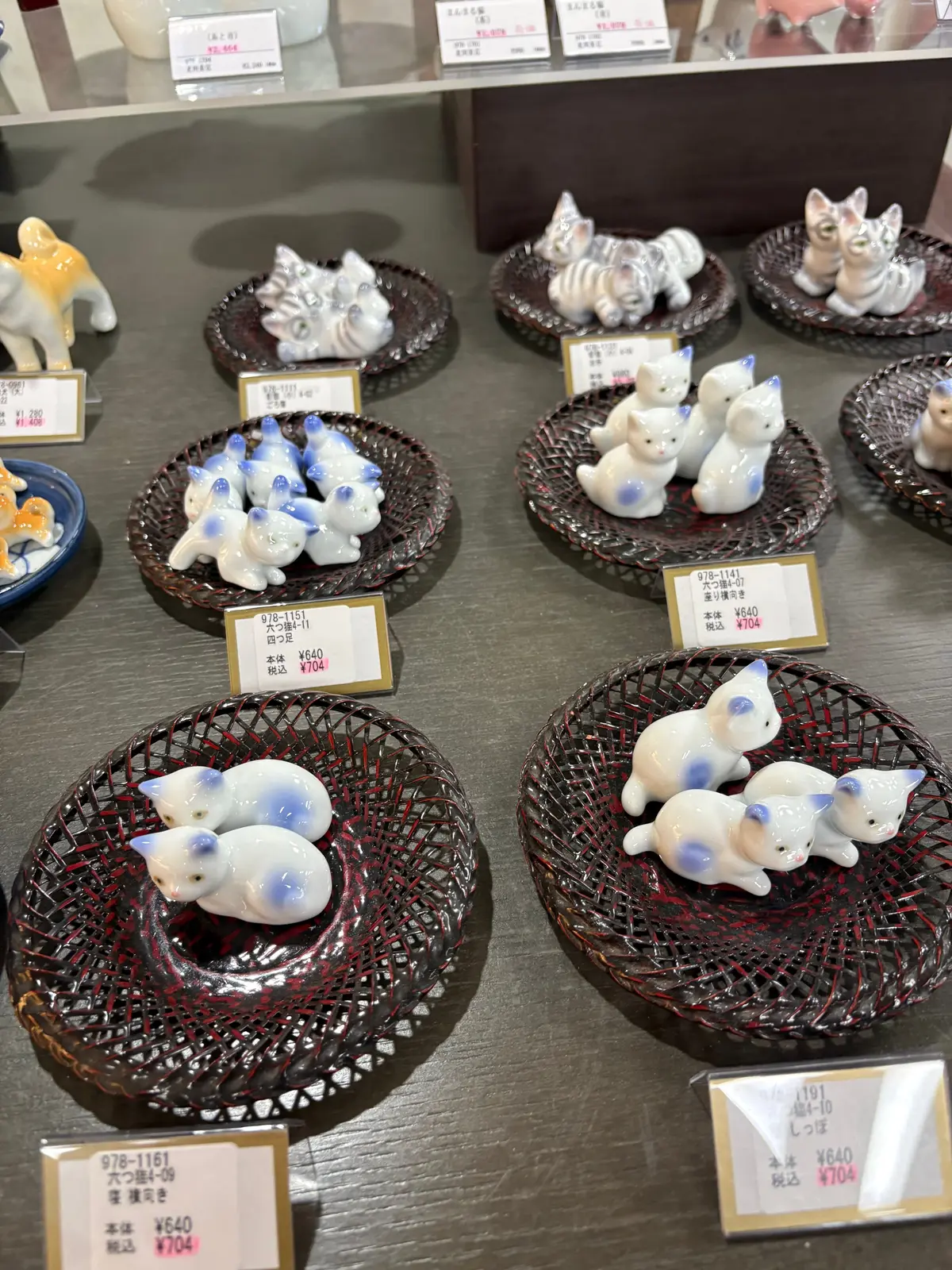 白と青の猫の置物が複数の編みバスケットに並べられ、価格タグが添えられている