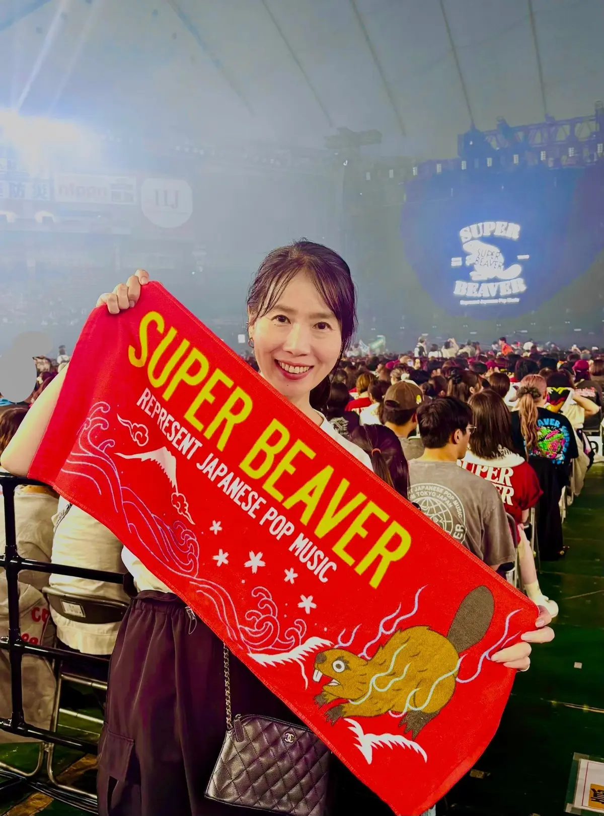 ライブ会場で、赤い「SUPER BEAVER」の応援棒を持っている女性。黒いワンピースにシルバーのバッグ、笑顔でこちらを見ている。背景にはステージと観客席が見える。