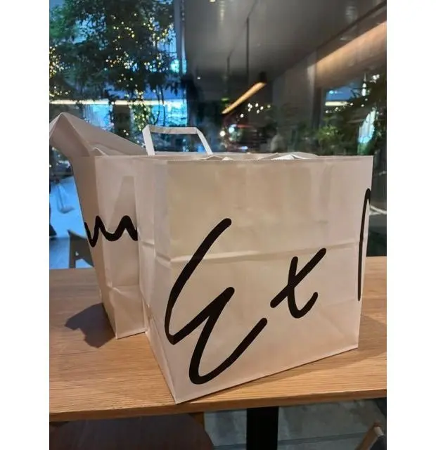 木製のテーブルの上に置かれた、白い紙袋2つ。片方の袋には黒い文字で「GX」と大きく書かれている。背景には、植物や光の反射が見える。