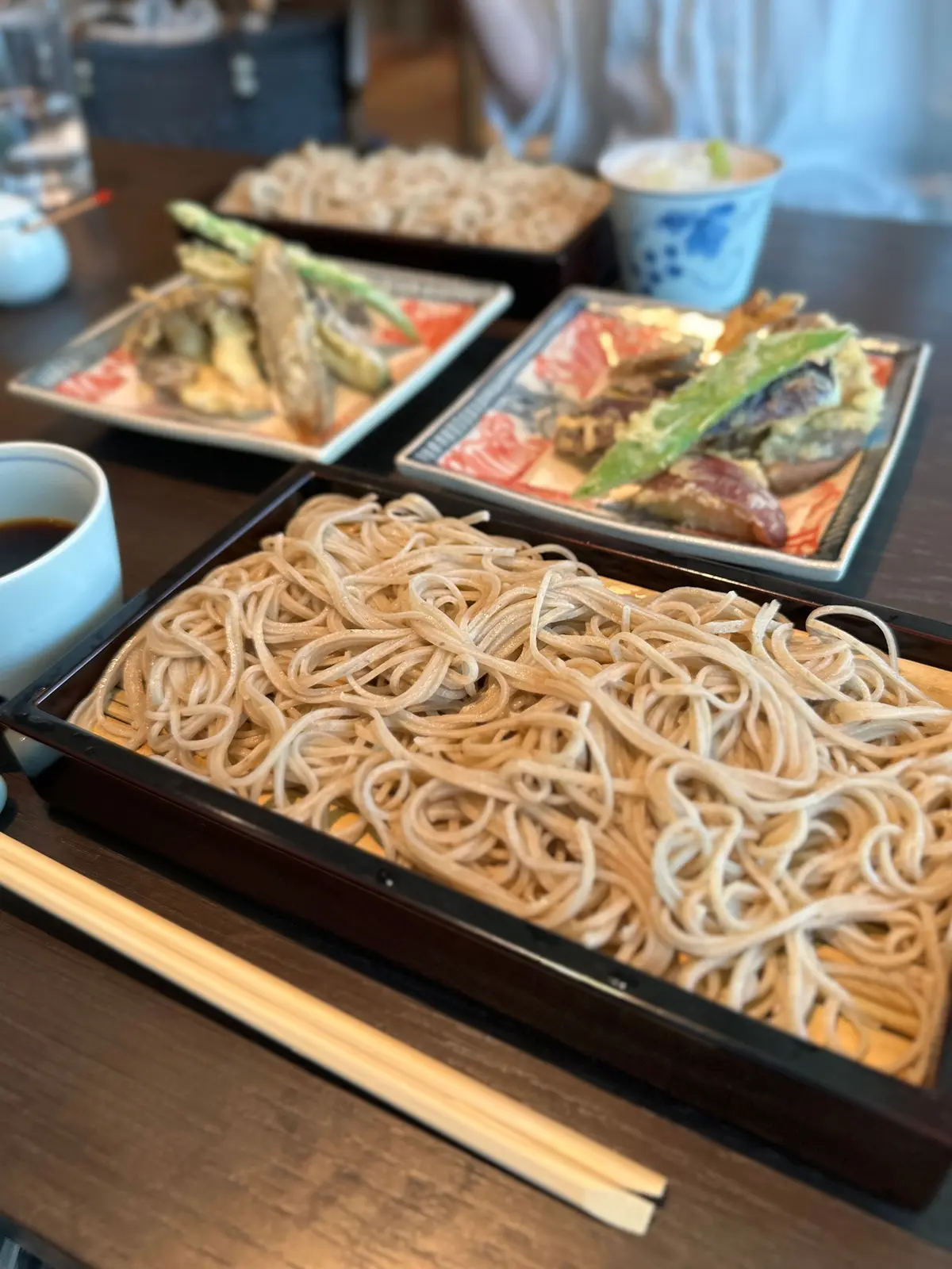茶色の木製テーブルに、うどんが盛られた黒い長方形の器があり、横に木製の箸が置かれている。周囲には刺身、焼き魚、揚げ物など他の料理が並び、青い花が飾られた白いカップとコーヒーも見える。