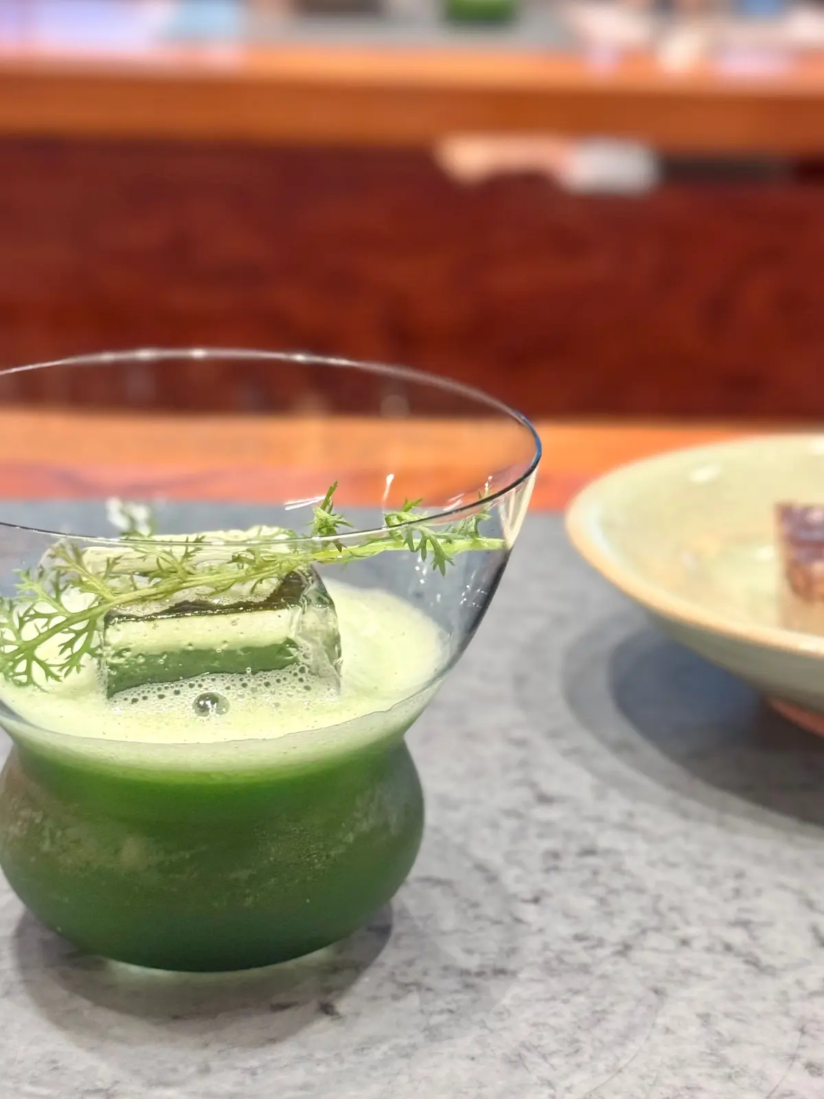 ガラスカップに緑色の泡立つ飲み物が入っており、ディルなどのハーブで飾られている。背景には木製のテーブルと別の料理が置かれている。