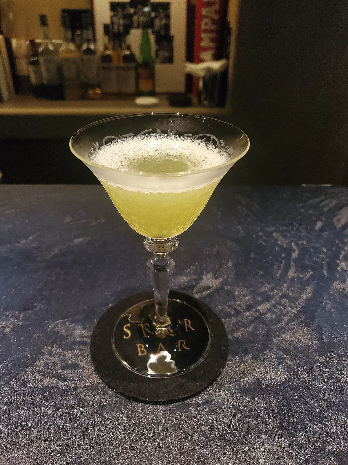 ガラスのカクテルグラスに黄色の泡立つ飲み物が入っている。グラスの台座には「STAR BAR」という文字が刻まれている。