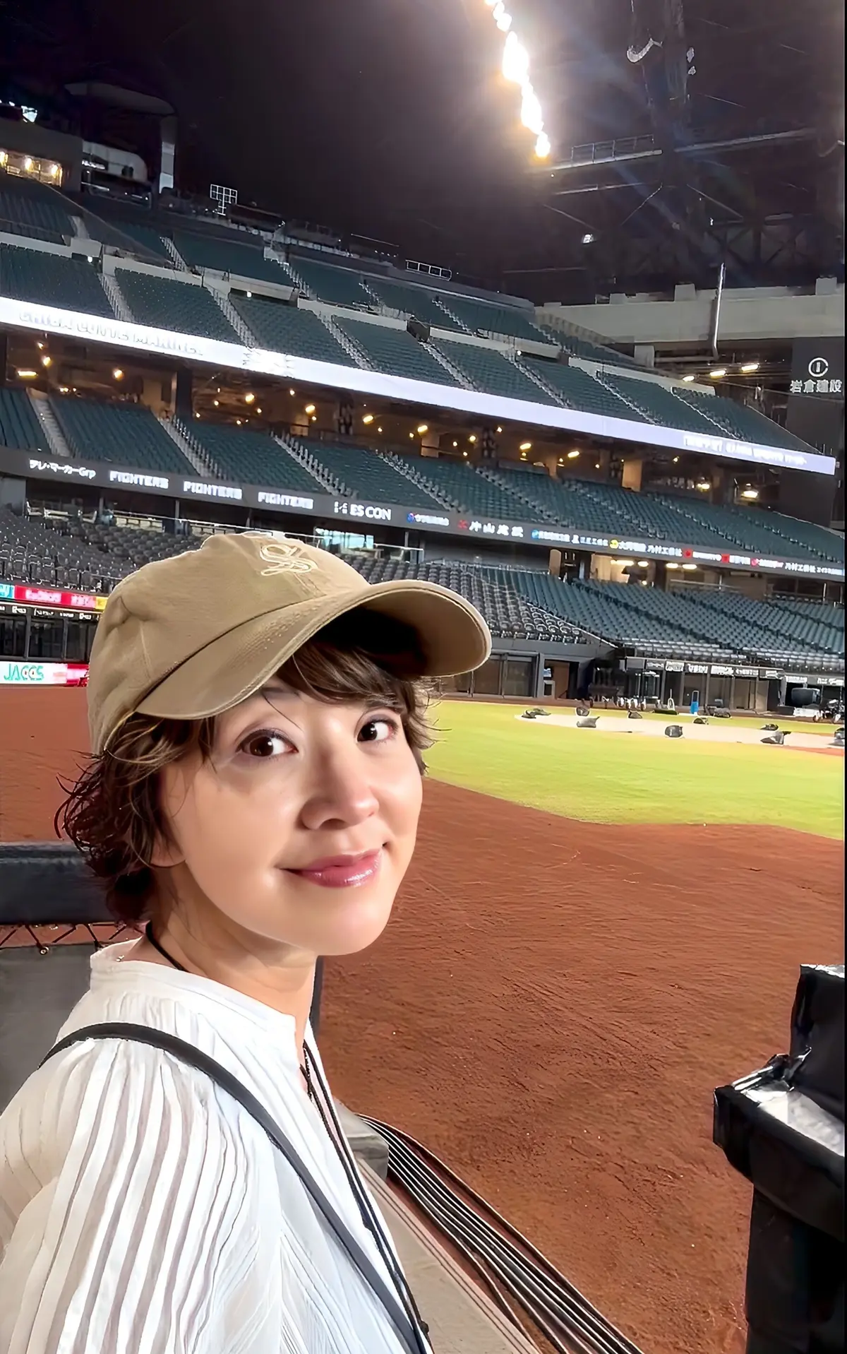 球場に立つ女性。茶色のキャップを被り、白いケーブルニットのトップスを着ている。背景には観客席とグラウンドが広がり、夜空には花火が上がっている。
