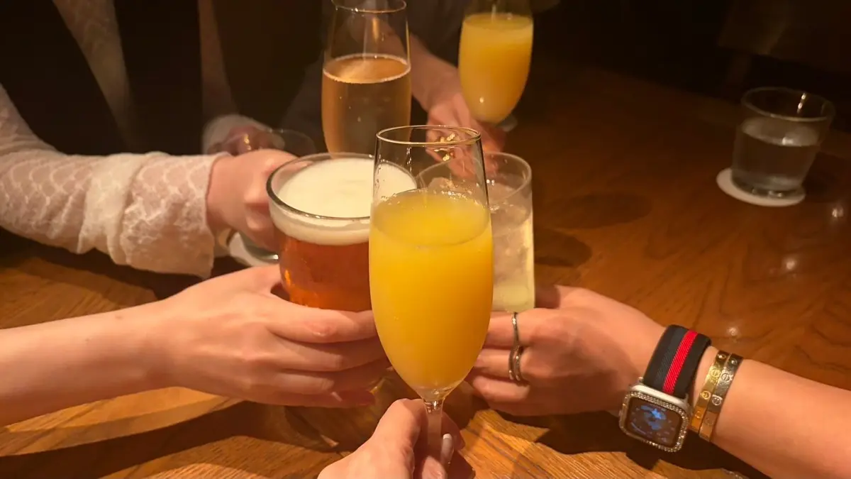 木製のテーブルを囲み、複数の人がグラスを手に合わせて乾杯している様子。オレンジ色のミモザ、ビール、透明な飲み物など、様々な色の液体が入ったグラスが並んでいる。