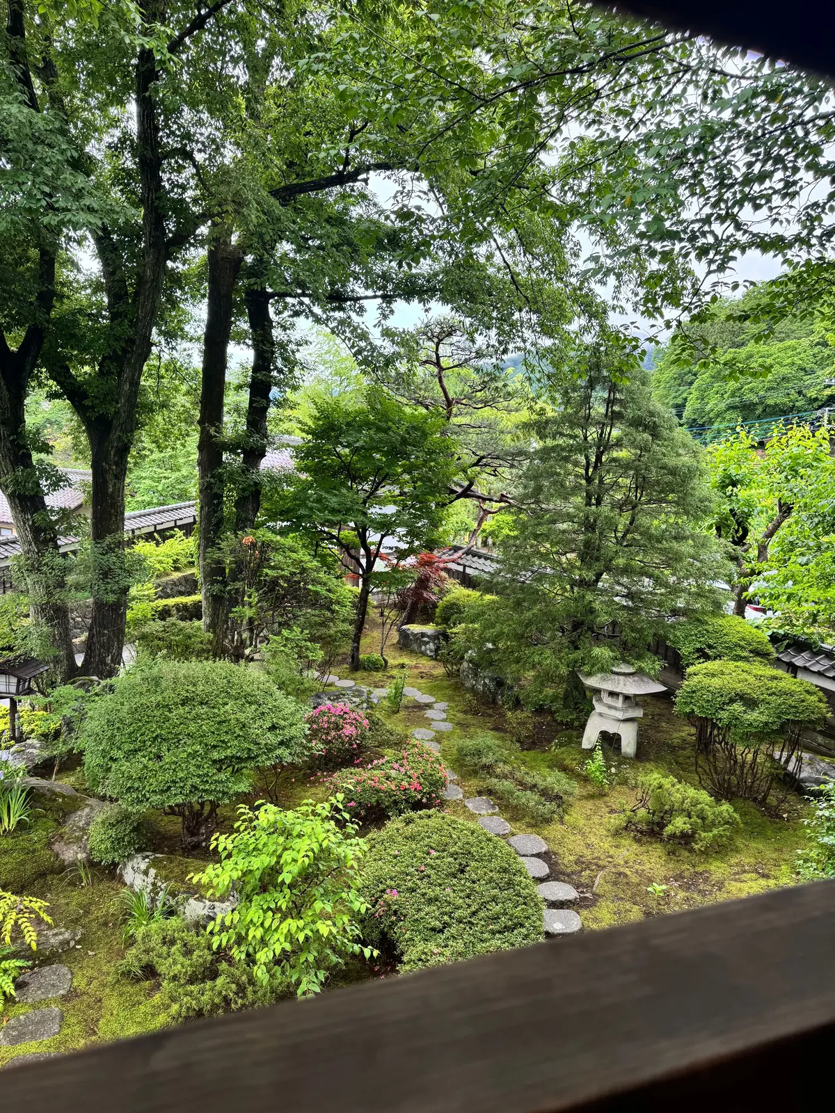 緑豊かな庭園。石畳の小道が中央を走り、左右に様々な樹木や草花が配置されている。中央には灯籠が置かれ、背景には伝統的な建築物が見える。