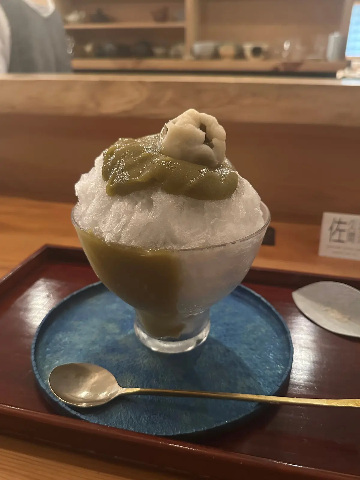 ガラスの器に入ったかき氷。緑色のシロップがかかり、上には白っぽい団子のようなものが乗っている。木製のトレイとスプーンが添えられている。背景には木製のカウンターと棚が見える。