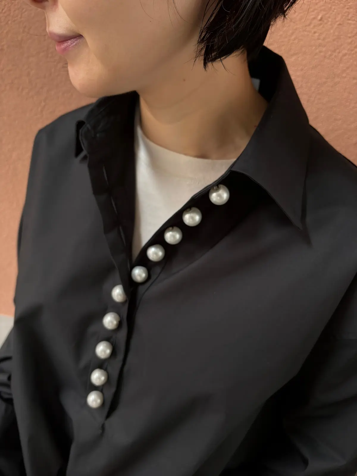 黒いシャツを着用した人物のアップ。白いTシャツを中に着ており、シャツの襟元には大小の真珠が並んでいる。背景は淡いオレンジ色。