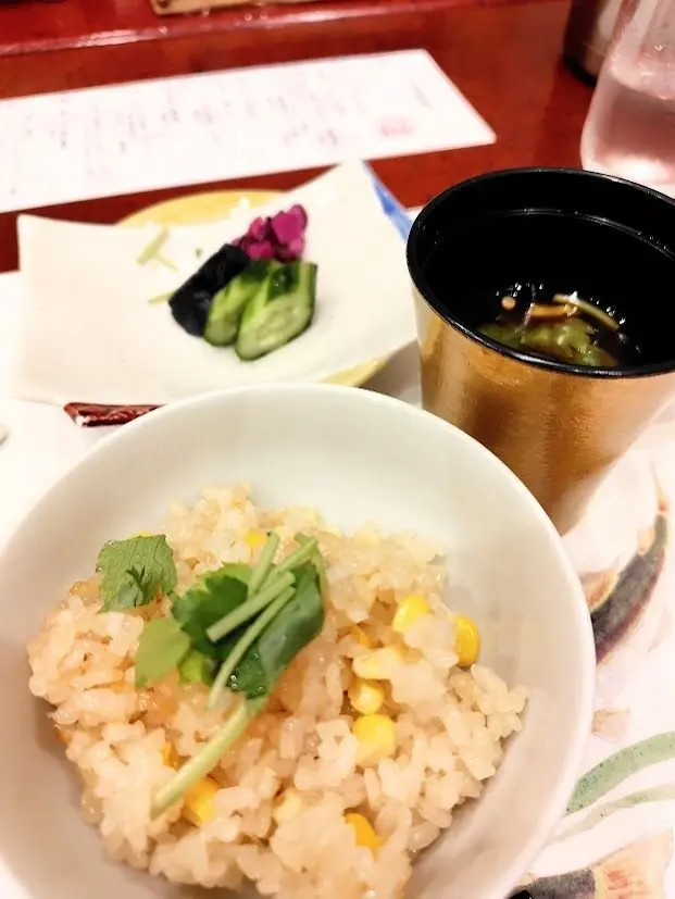 白いボウルにコーン入りご飯とパクチーの飾りが入っており、隣にはキュウリと花の飾りが乗ったプレートと黒いカップに緑の汁物がある