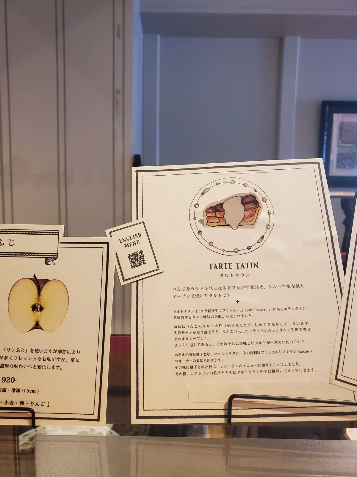 カフェのメニューの一部。右ページには「TARTE TATIN（タルトタタン）」のイラストと説明が記載。タルトタタンの断面図が描かれ、りんごやキャラメル色。左ページには他のメニューのイラストと日本語のテキスト。QRコードも掲載。