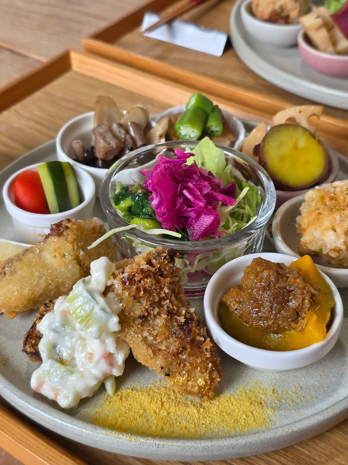 木製のトレイに盛り付けられた、様々な小鉢料理。中央には、ピンク色のキャベツと葉物野菜のサラダ。周りには、魚のフライ、和え物、漬物、黄色い粉末などが彩り豊かに並んでいる。