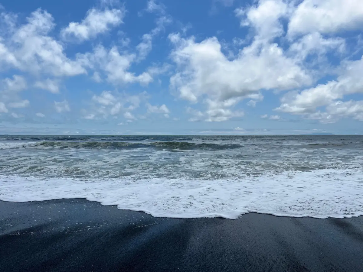 青空と白い雲が広がる海、波が黒い砂浜に打ち寄せる風景