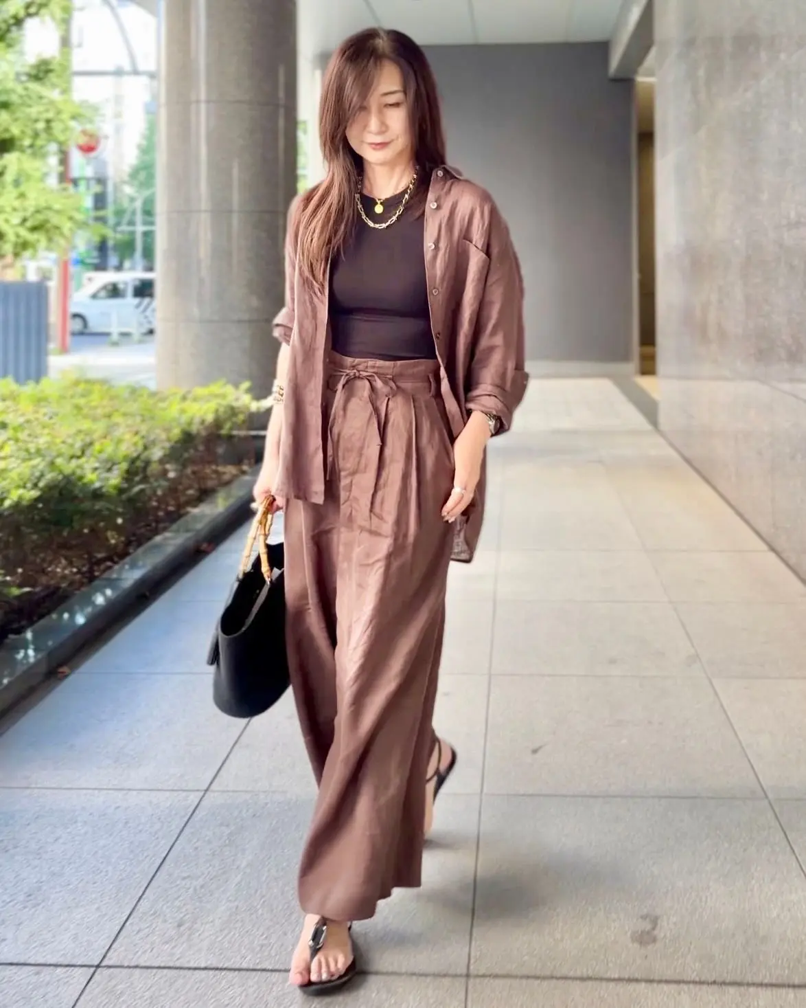 ブラウンのシャツとワイドパンツのセットアップを着た女性が歩いている全身写真。黒のタンクトップを合わせ、ゴールドのネックレスを着用。黒いバッグを持ち、黒いサンダルを履いている。背景はコンクリートの建物と植栽。