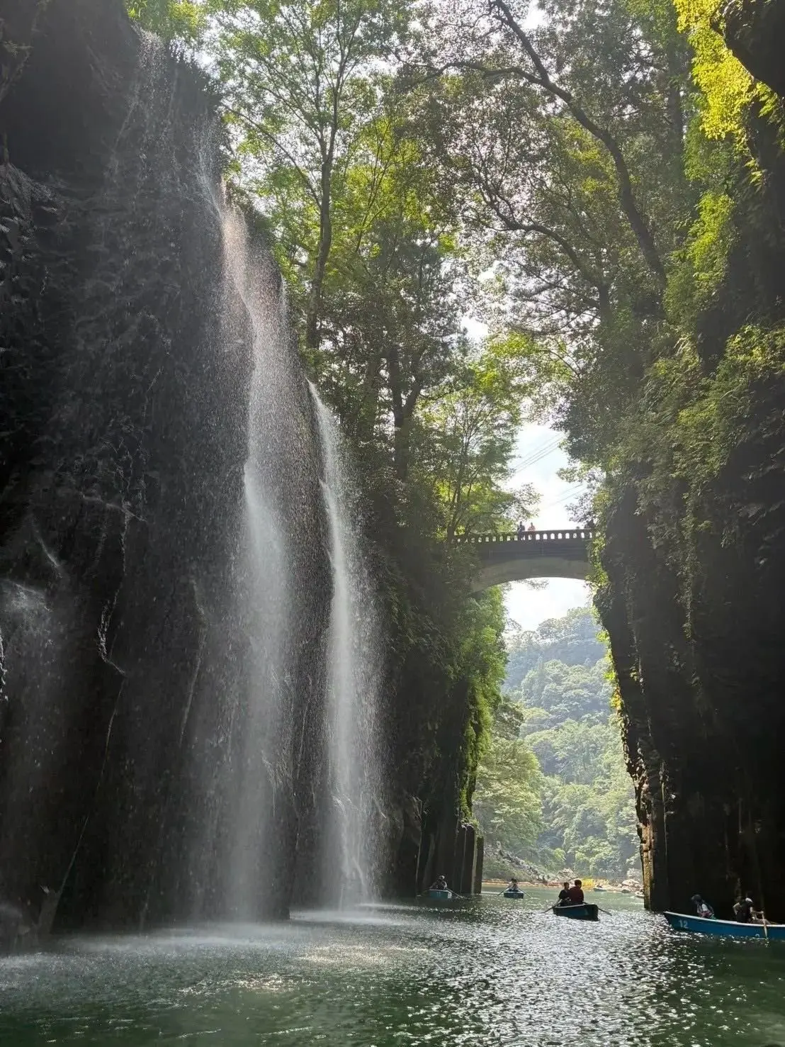 深い峡谷に流れ落ちる滝と、水面を漂うボート。緑豊かな木々が生い茂り、遠くには橋が見える。
