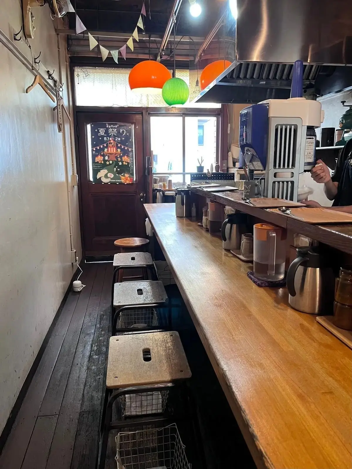 木製のカウンターと椅子が並ぶ、日本の居酒屋のような内装。左にはメニューらしきポスター、右には調理器具や食器が置かれている。天井には緑色のペーパーランタンが吊るされている。