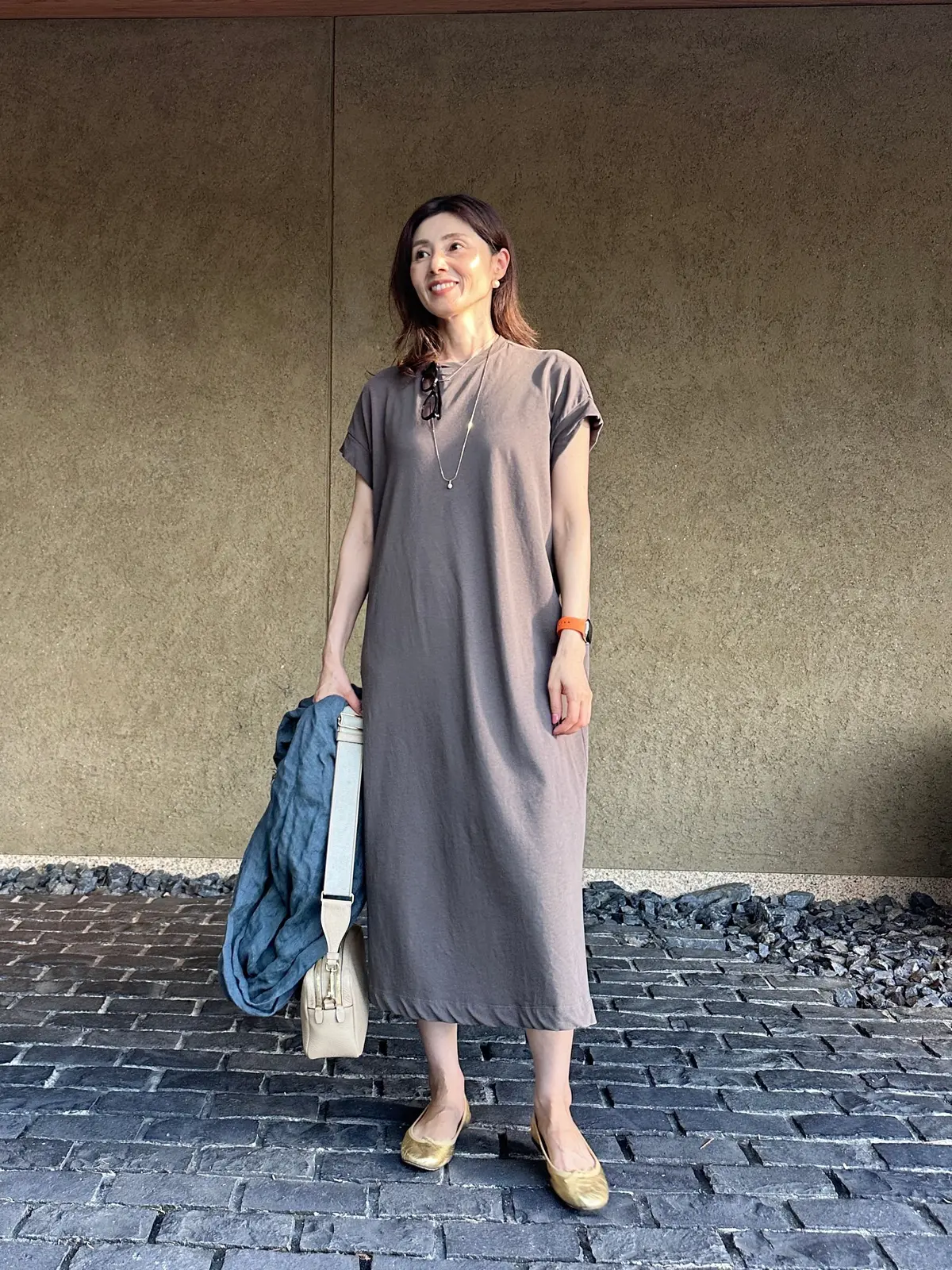 女性がグレーのロングドレスを着て、コンクリート壁の前で立っている。左手には青いジャケットと白いトートバッグを保持し、右手にはオレンジのアクセサリーが見える。足元はベージュのフラットシューズ。