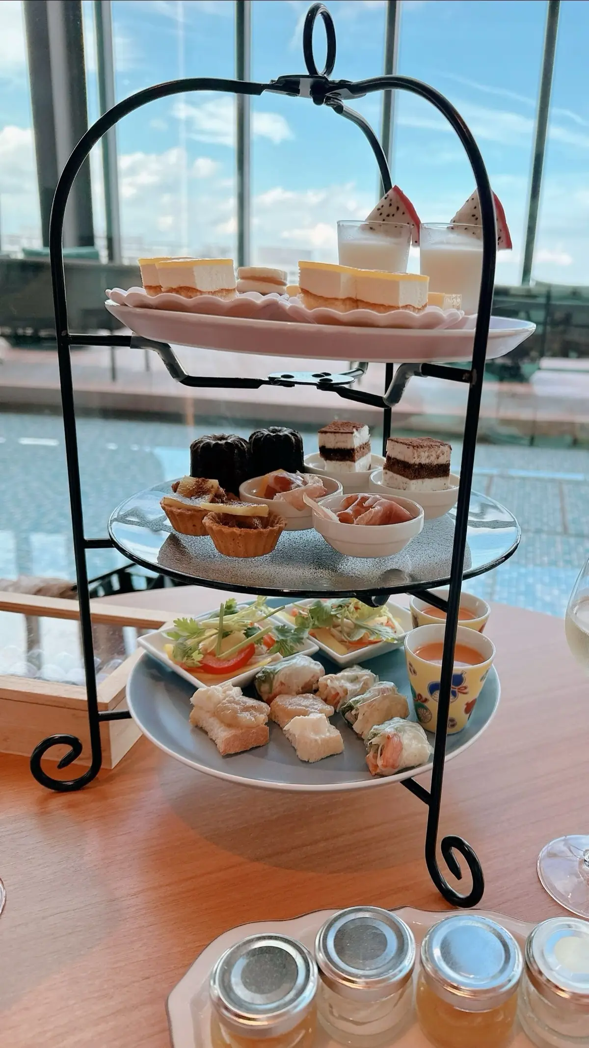 三段のケーキスタンドに、軽食とスイーツが盛り付けられている。一番下はサンドイッチや春巻き、真ん中にはタルトやチョコレート、一番上にはチーズケーキやフルーツのデザート。背景には窓から見える青空とプールが見える。