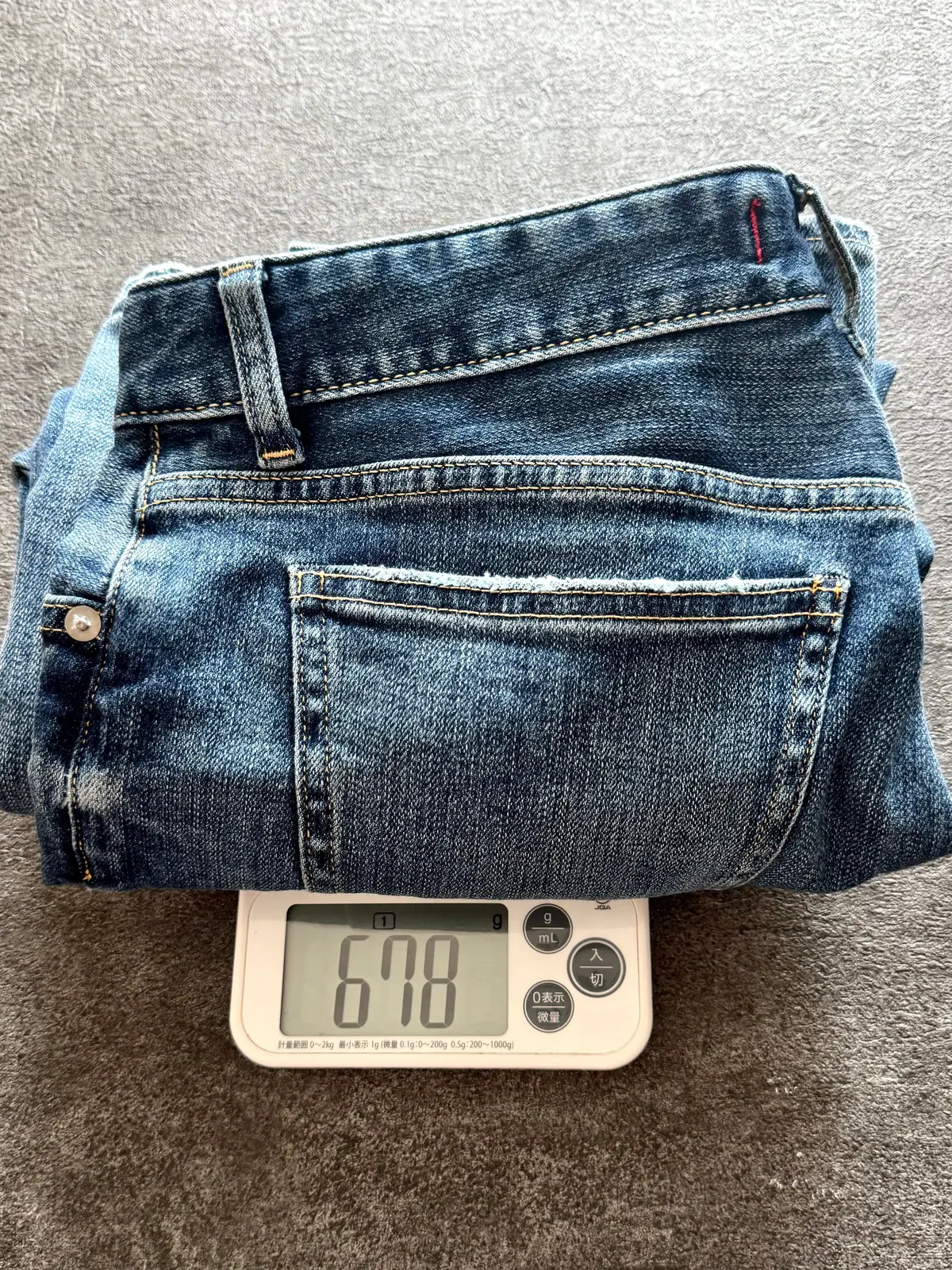 デニムパンツが電子秤の上に置かれ、重さが678グラムと表示されている