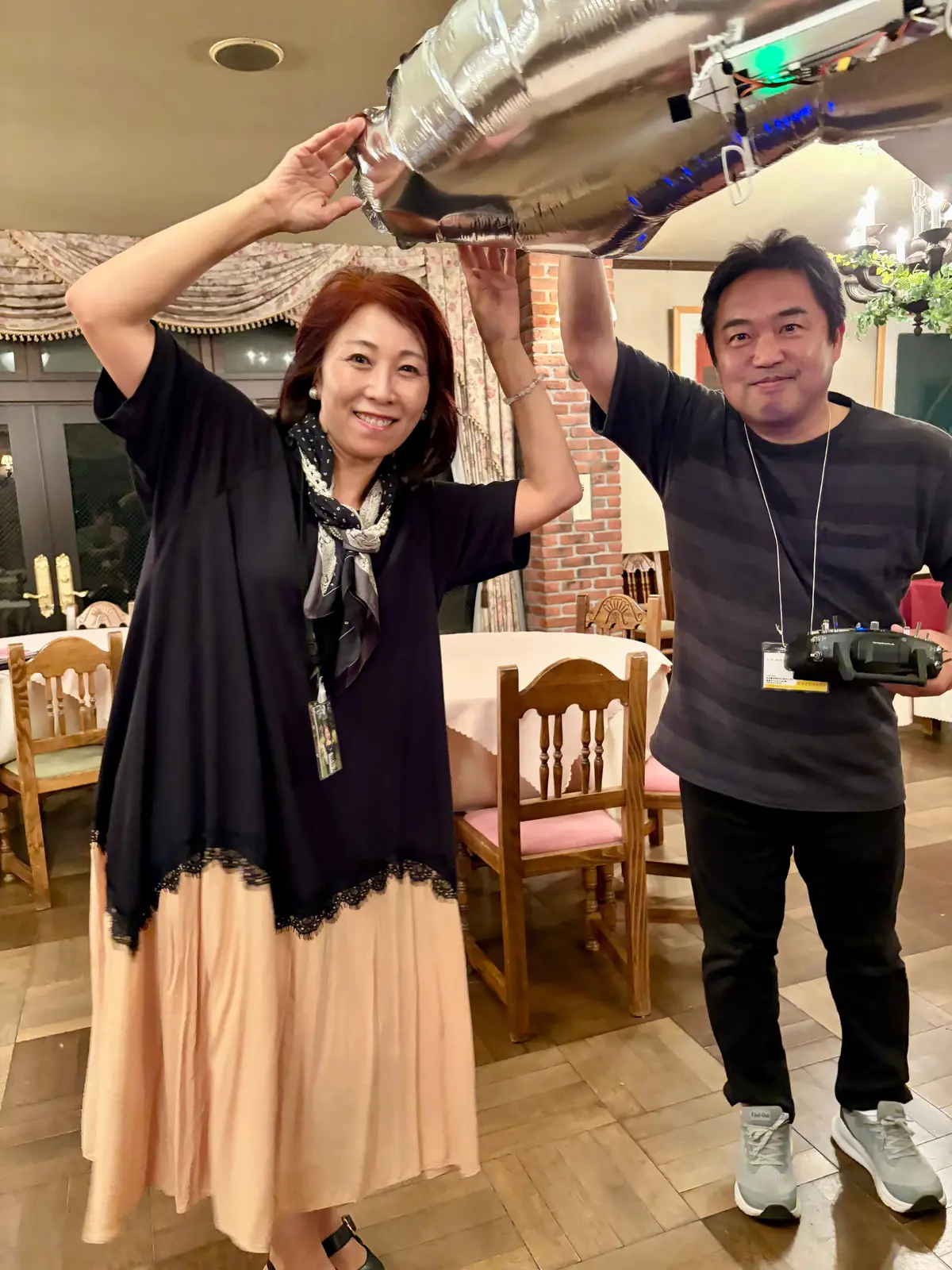 笑顔の女性と男性が、銀色の風船を持ち上げて喜んでいる。背景にはテーブルや椅子が並び、温かい照明が灯っている。