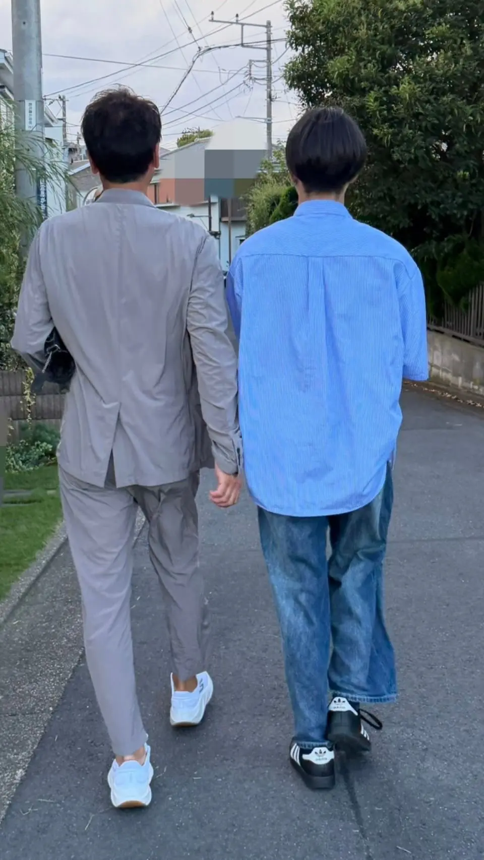 2人の男性が路肩を寄り添って歩いている後ろ姿。左はグレーのスーツ、右は青いシャツとジーンズ姿。左は白いスニーカー、右は黒いAdidasのスニーカーを履いている。背景には電線と緑の木々が見える。