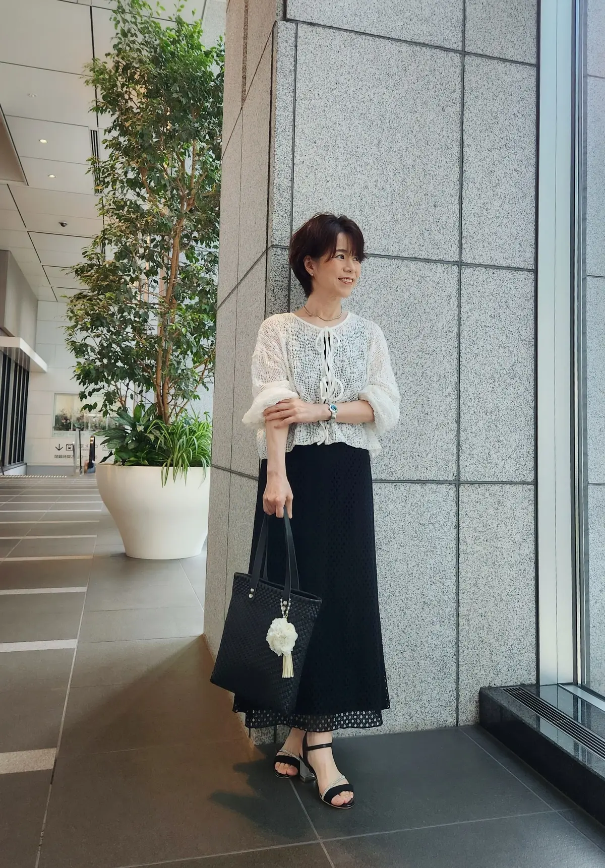 女性が白い編み物トップスと黒いスカートを着用し、黒いトートバッグを手に持って、モダンな建物の壁に寄りかかっている。背景には大きな観葉植物とガラスの窓がある。