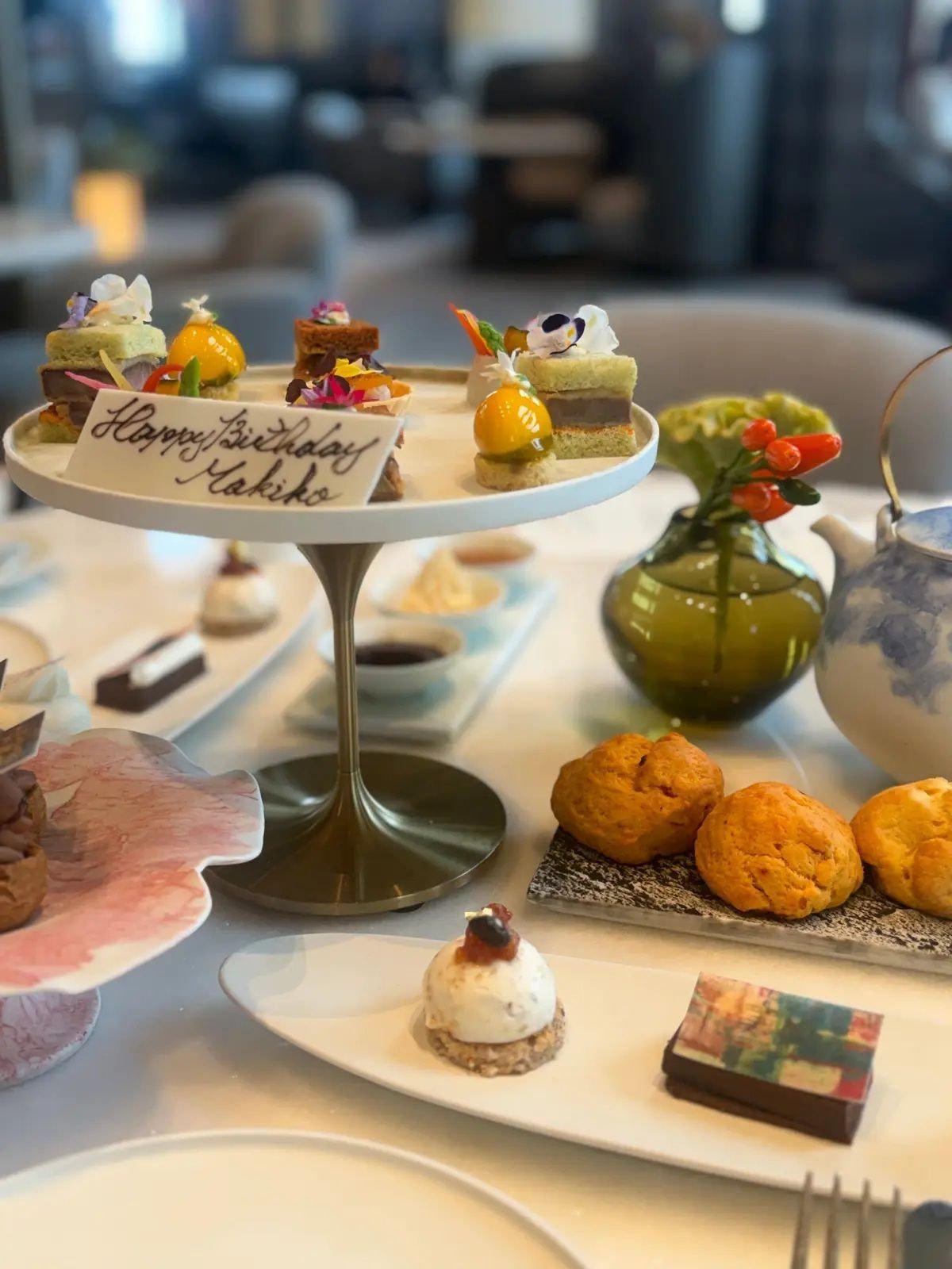 白テーブルに置かれた、誕生日のお祝いの Afternoon Tea セット。2段のスタンドに様々なミニケーキや焼き菓子、スコーンが並び、緑色のデミタッセや花瓶が添えられている。