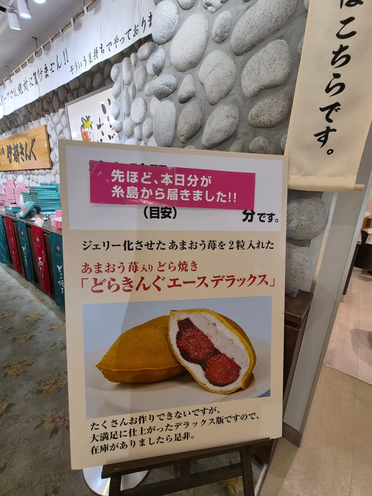 お店のメニューボード。ピンク色の枠に「先ほど、本日から糸島から届きました！」と記載。ジェリー化されたあまおり2枚入りの「あまおり入り どら焼き「どらきんぐ」」の写真が中央にあり、横にはチップとポテト。背景には、石積み風の装飾と商品が並ぶ棚。