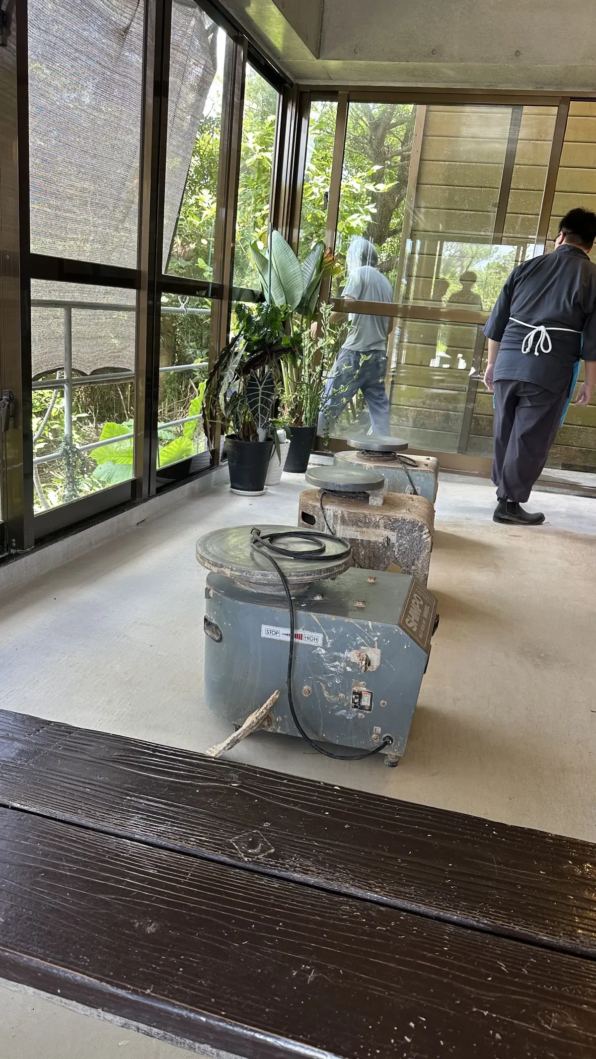 ガラス張りの空間に、陶芸の轆轤（ろくろ）が置かれている。中央には青い轆轤があり、その周りには植木鉢に入った植物が並んでいる。背景には緑豊かな自然が見え、右には黒い制服を着た男性が立っている。