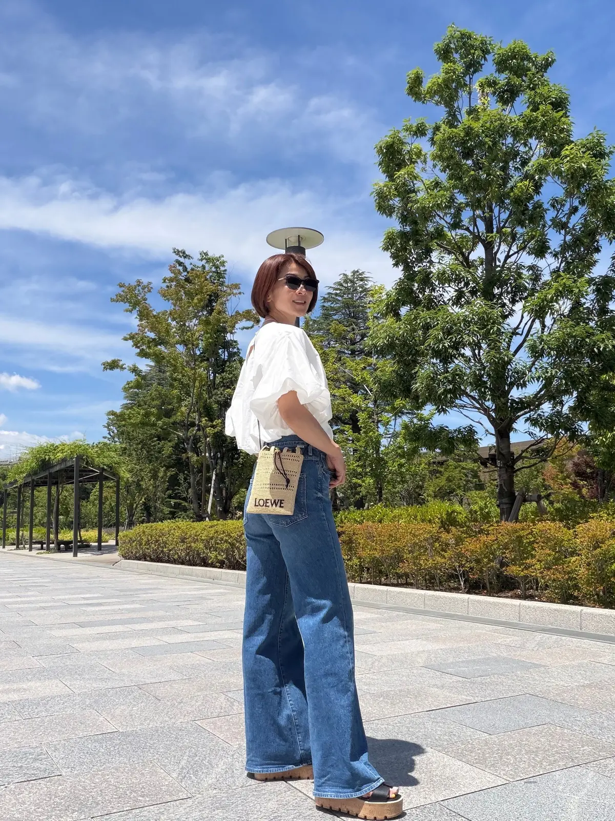 白いブラウスとデニムパンツの女性が、木々のある公園でサングラスをかけ、LOEWEのトートバッグを肩にかけ、晴れた空の下で立っている。