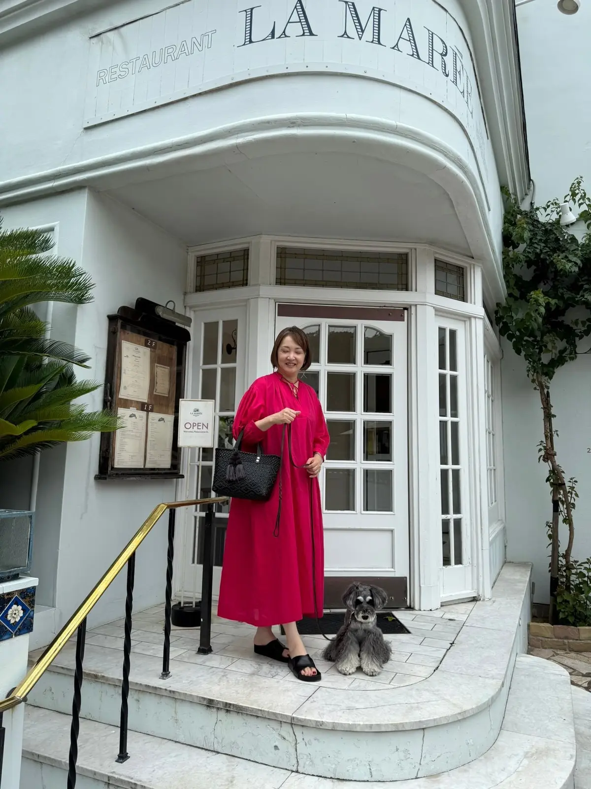 ピンクのワンピースを着た女性と、灰色の毛並みの犬がレストランの入り口に立つ。白とグレーのストライプ模様のサンダルを履き、黒いカバンを持つ。背景には「LA MARE」と書かれたレストランの看板が見える。