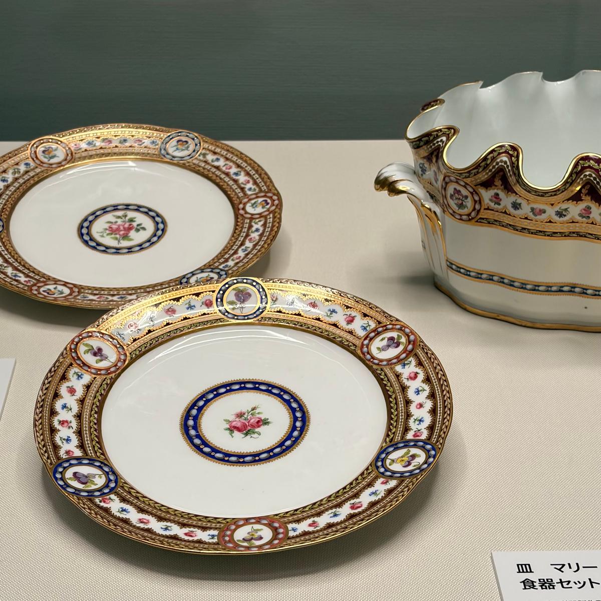 金彩と花模様が施された磁器製の皿2枚と、同じデザインの湯呑み。皿は白地に金色の縁取り、中央に花が描かれている。湯呑みは白地に金彩の装飾が施され、柄は皿と共通。