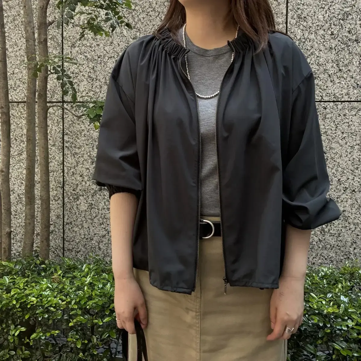 黒のジャケットとベージュのスカートを着た女性が、石造りの壁と緑の低木の前で立っています。