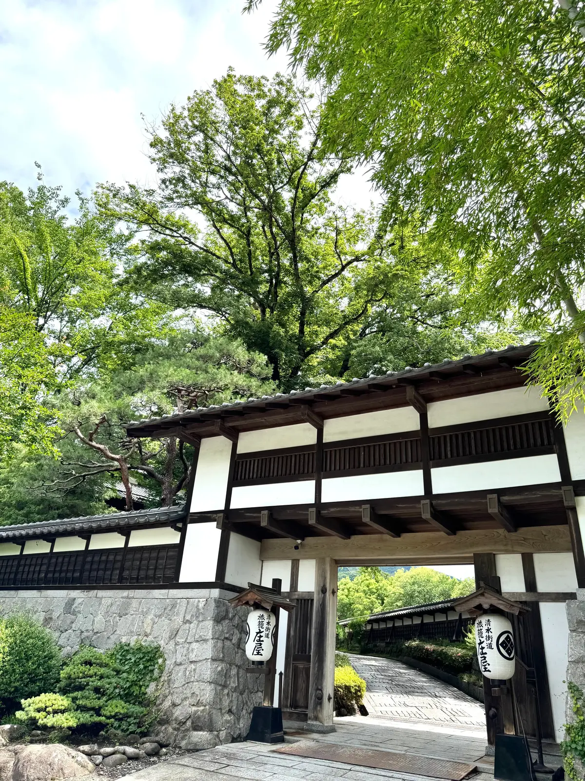 木々に囲まれた伝統的な日本家屋の門。白漆喰の壁と木製の柱、瓦屋根が特徴的。左右には提灯のようなものがあり、漢字の文字が書かれている。石畳の道が奥へと続いている。