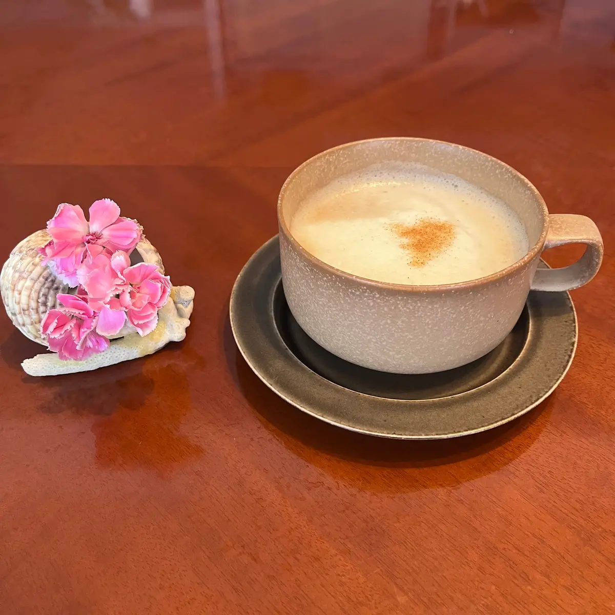 茶色のカップとソーサーに注がれたラテ。表面にはシナモンでハートの模様が描かれている。隣にはピンク色の花が飾られた貝殻が置かれている。