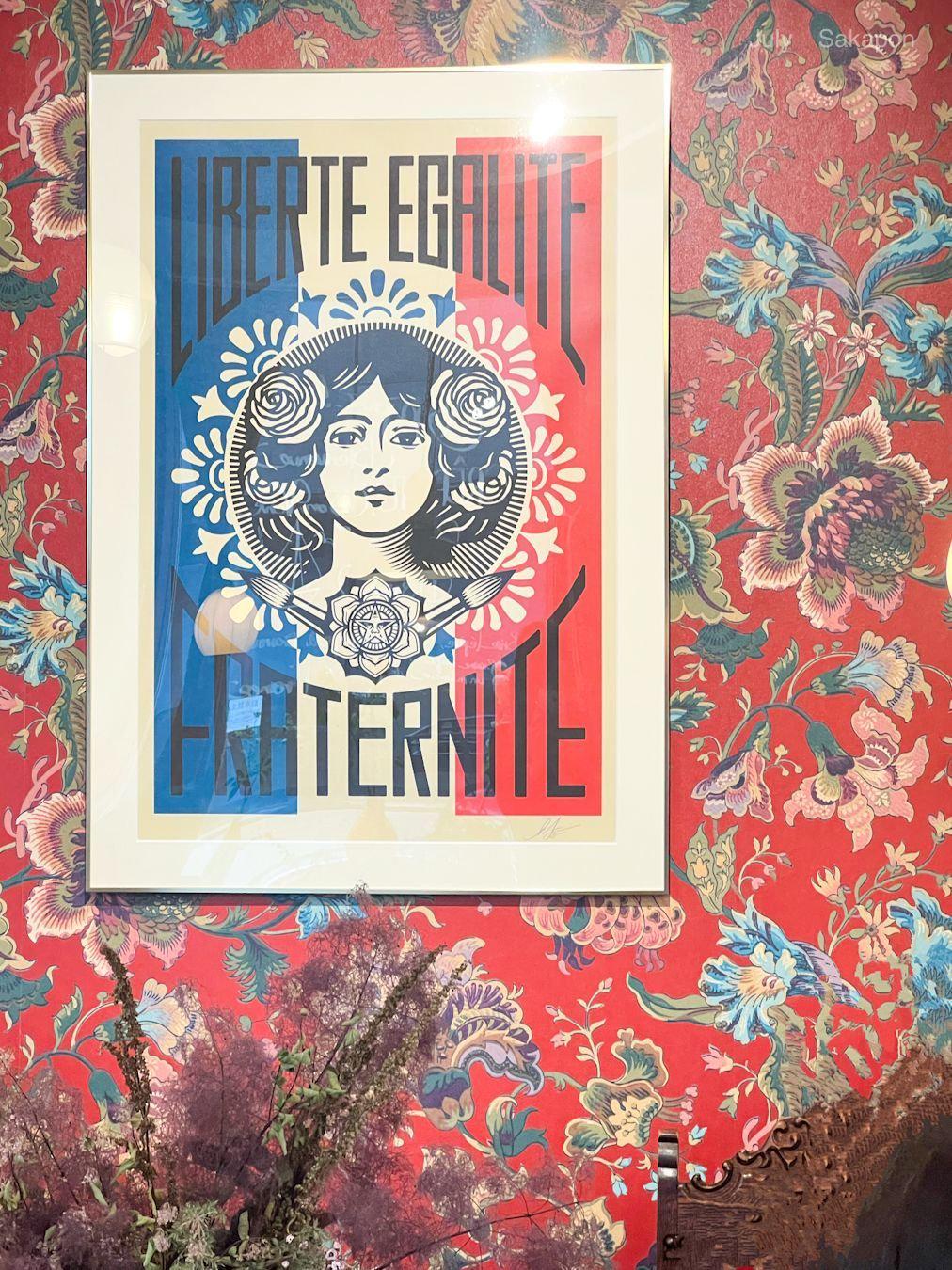 赤い花模様の壁に額装されたポスター。女性像が中央に描かれ、頭上に「LIBERTE EGALITE」と「FRATERNITE」の文字が青と赤で表示されている。