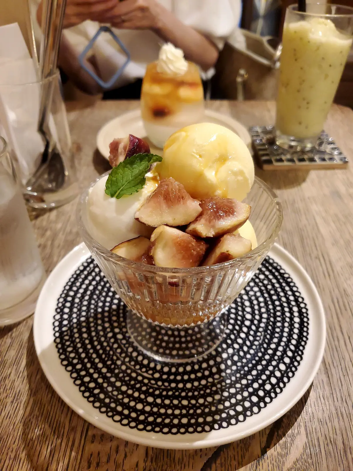 ガラスの器に入ったデザート。ヨーグルト、焼きフィグ、バニラアイスクリーム、ミントの葉が盛り付けられている。黒と白の模様の皿の上に置かれ、背景には飲み物が写っている。
