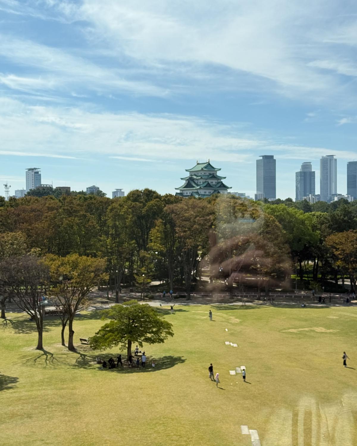 大阪城天守閣を中心に、緑豊かな公園と近代的な高層ビル群が広がる風景。中央に天守閣、手前には人々が散策する芝生エリア、背景には高層ビルが並ぶ。