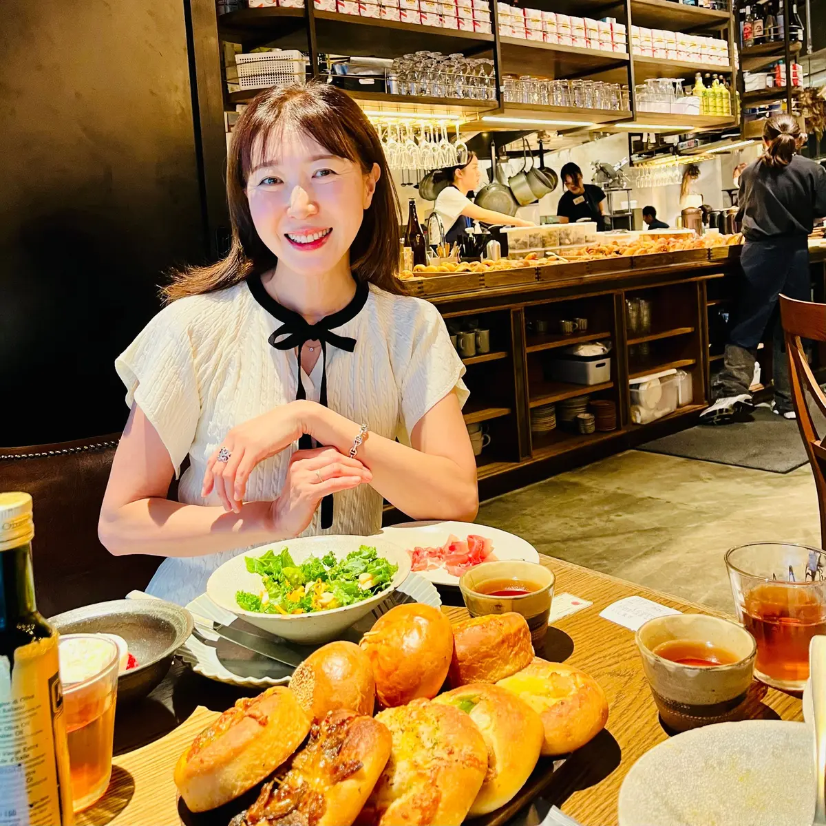女性がレストランのテーブルに座り、笑顔でパンの盛り合わせ、サラダ、飲み物を前にしています。背景には調理場があり、スタッフが調理しています。